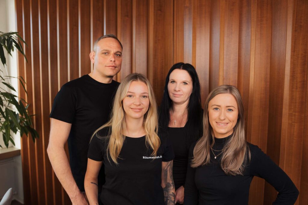 Vier Personen, drei Frauen und ein Mann, stehen gemeinsam in einem Innenraum vor einer holzgetäfelten Wand. Sie tragen schwarze Hemden und lächeln in die Kamera. Sie sehen aus, als wären sie bereit, bei der Räumung oder Altmöbelentsorgung zu helfen.