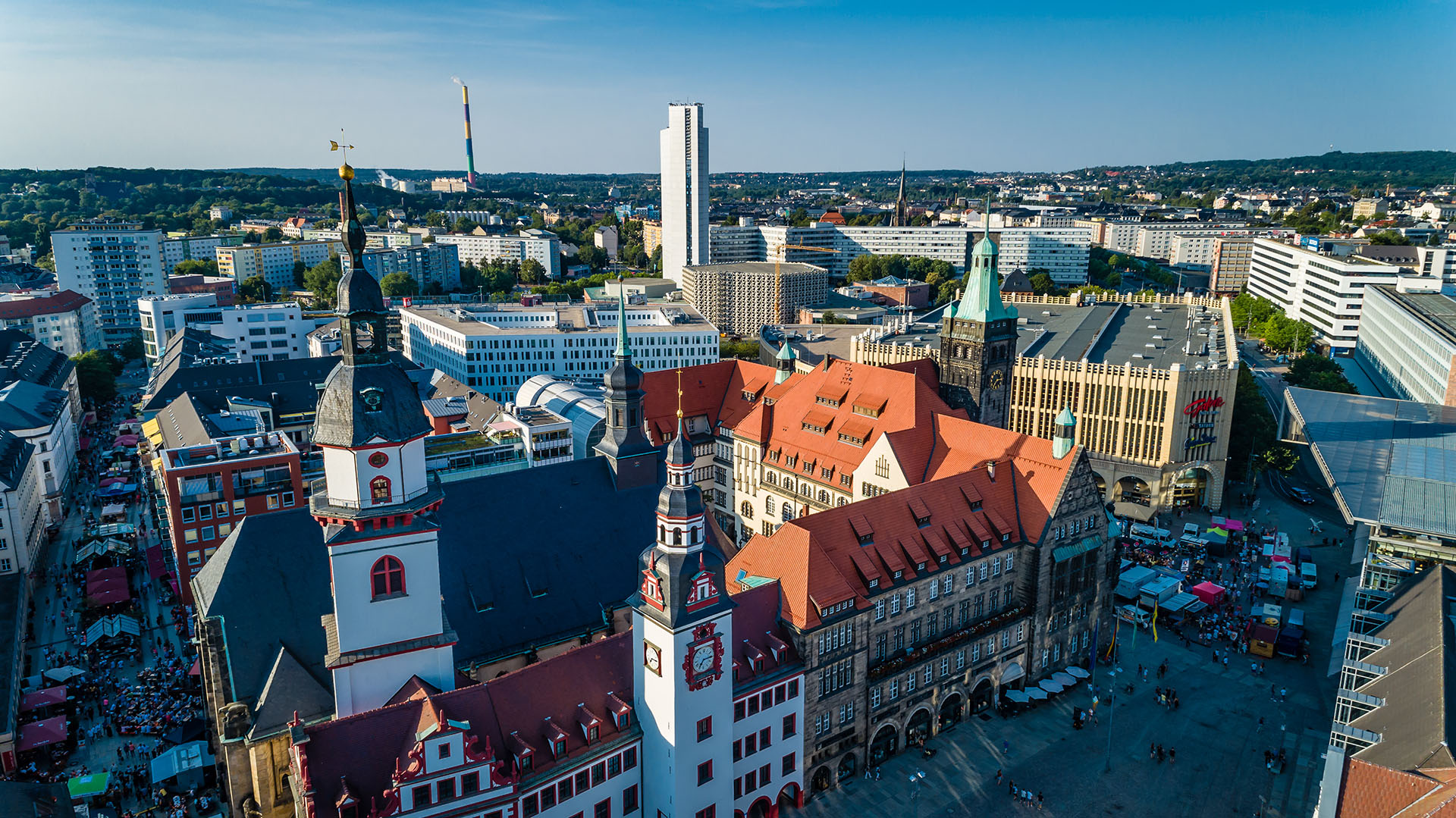 Luftaufnahme eines Stadtplatzes mit historischen Gebäuden, roten Ziegeldächern und Uhrentürmen; moderne Hochhäuser umgeben das Gebiet. Dienstleistungen wie Wohnungsauflösung und Altmöbelentsorgung tragen dazu bei, dass das Stadtbild unter dem blauen Himmel lebendig bleibt.