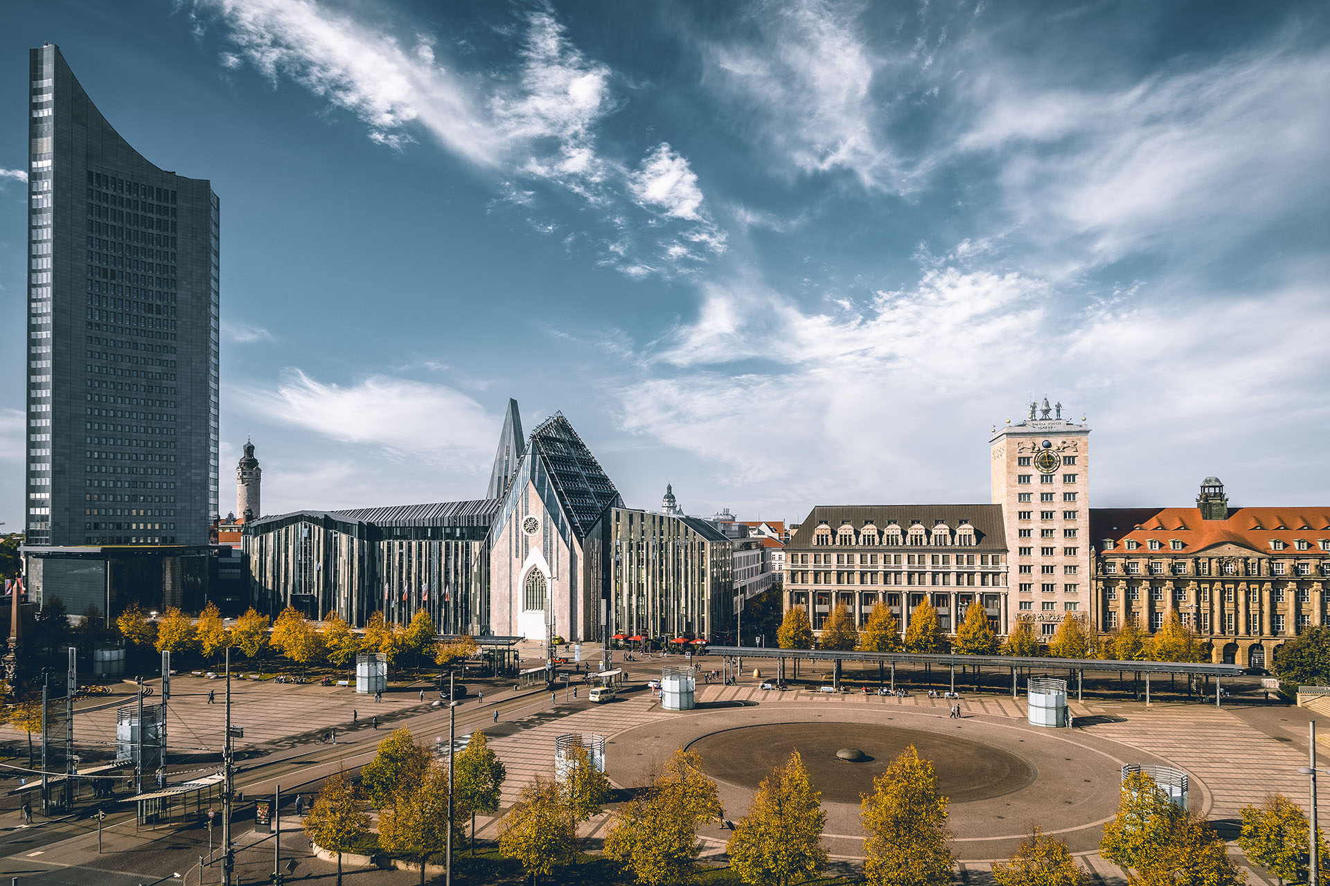 Ein breiter Stadtplatz mit modernen und historischen Gebäuden, darunter ein hoher gläserner Wolkenkratzer, ein kirchenähnlicher Bau mit Glasfassaden und ältere klassische Gebäude, umgeben von Bäumen unter blauem Himmel - ideal für die Entrümpelung oder Altmöbelentsorgung.