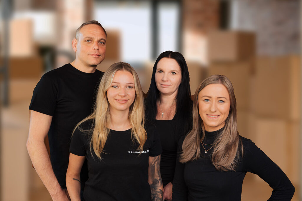 Vier Erwachsene - zwei Männer und zwei Frauen - stehen zusammen in einem Haus und lächeln in schwarzen Hemden in die Kamera. Im Hintergrund ist ein Raum mit gestapelten Pappkartons zu sehen, die auf eine Haushaltsauflösung in Dresden hindeuten.